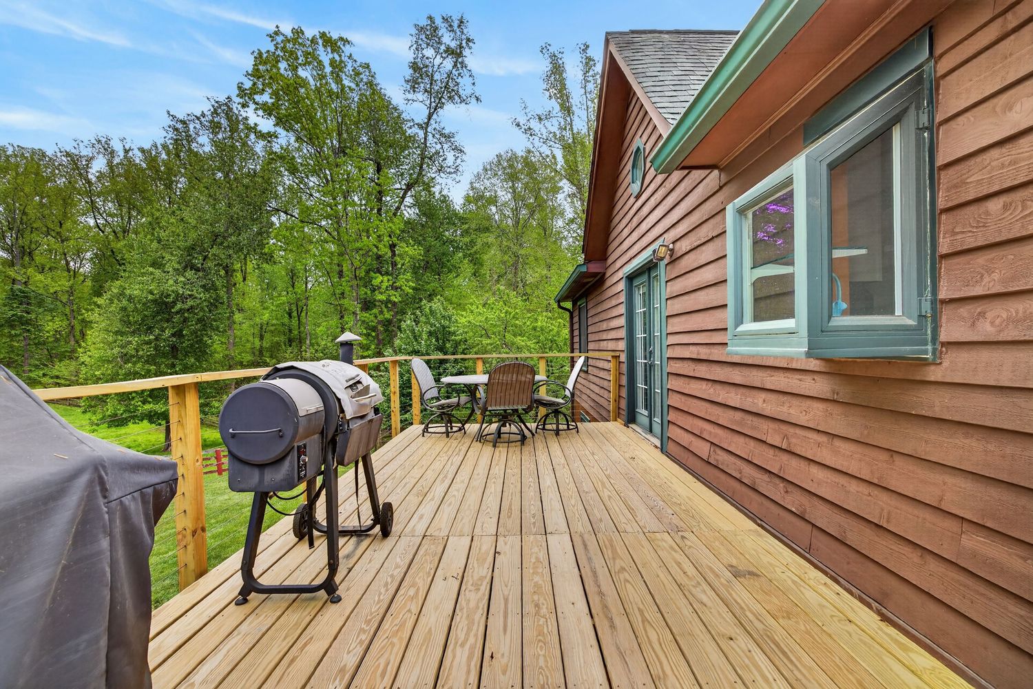 Wooden deck with BBQ grill and outdoor dining set, surrounded by lush green trees beside a rustic cabin.