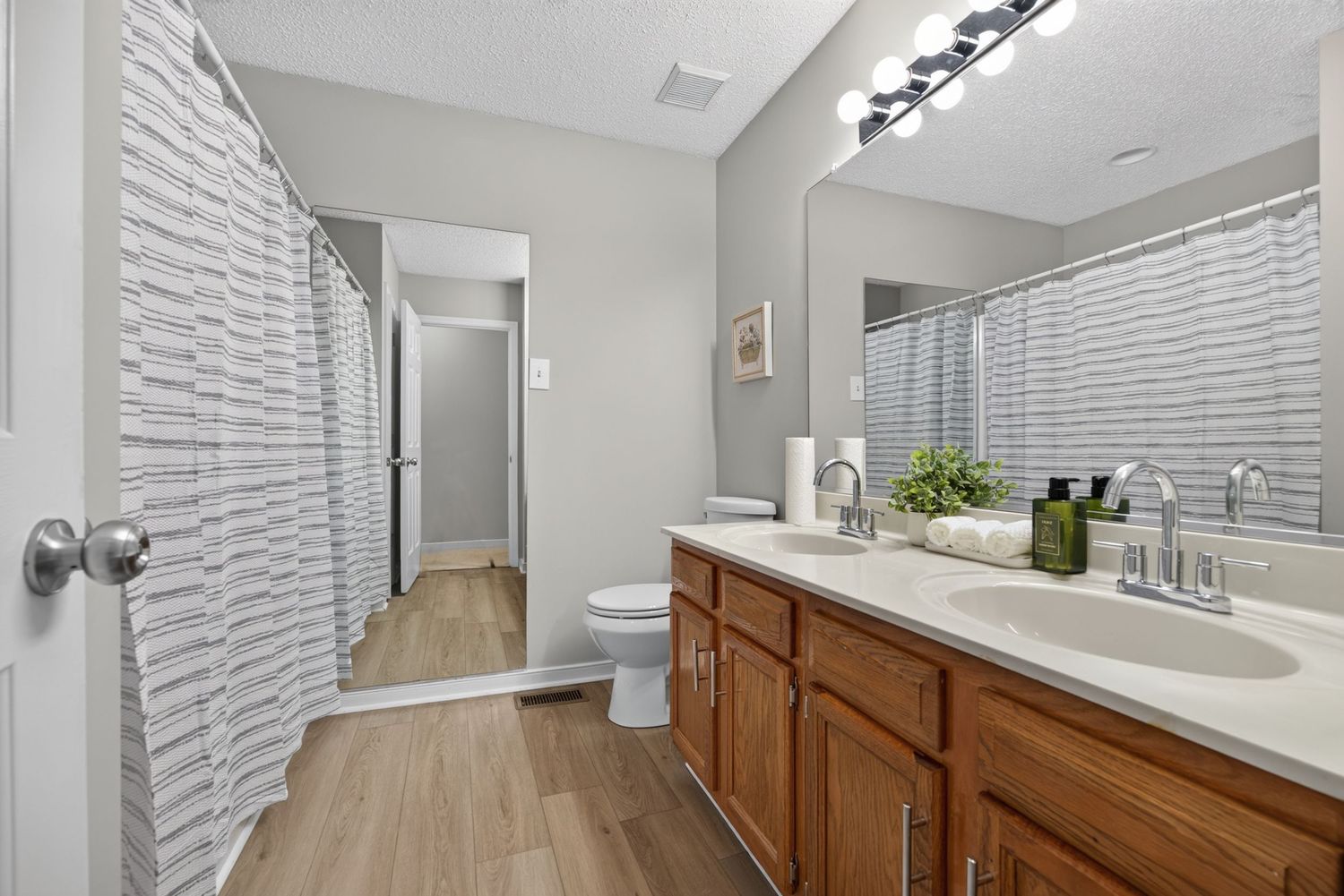 Spacious bathroom with double sink vanity, wood cabinets, large mirror, hardwood floors, and white shower curtain.