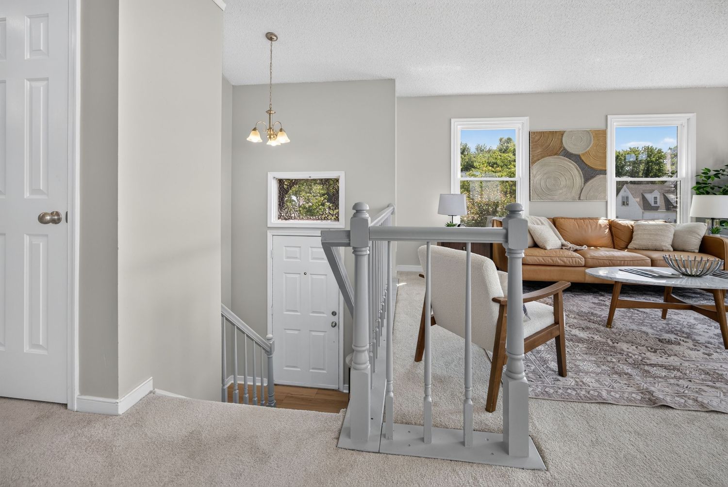 Bright living room with tan leather sofa, wood coffee table, gray stair railing, and large windows letting in natural light.