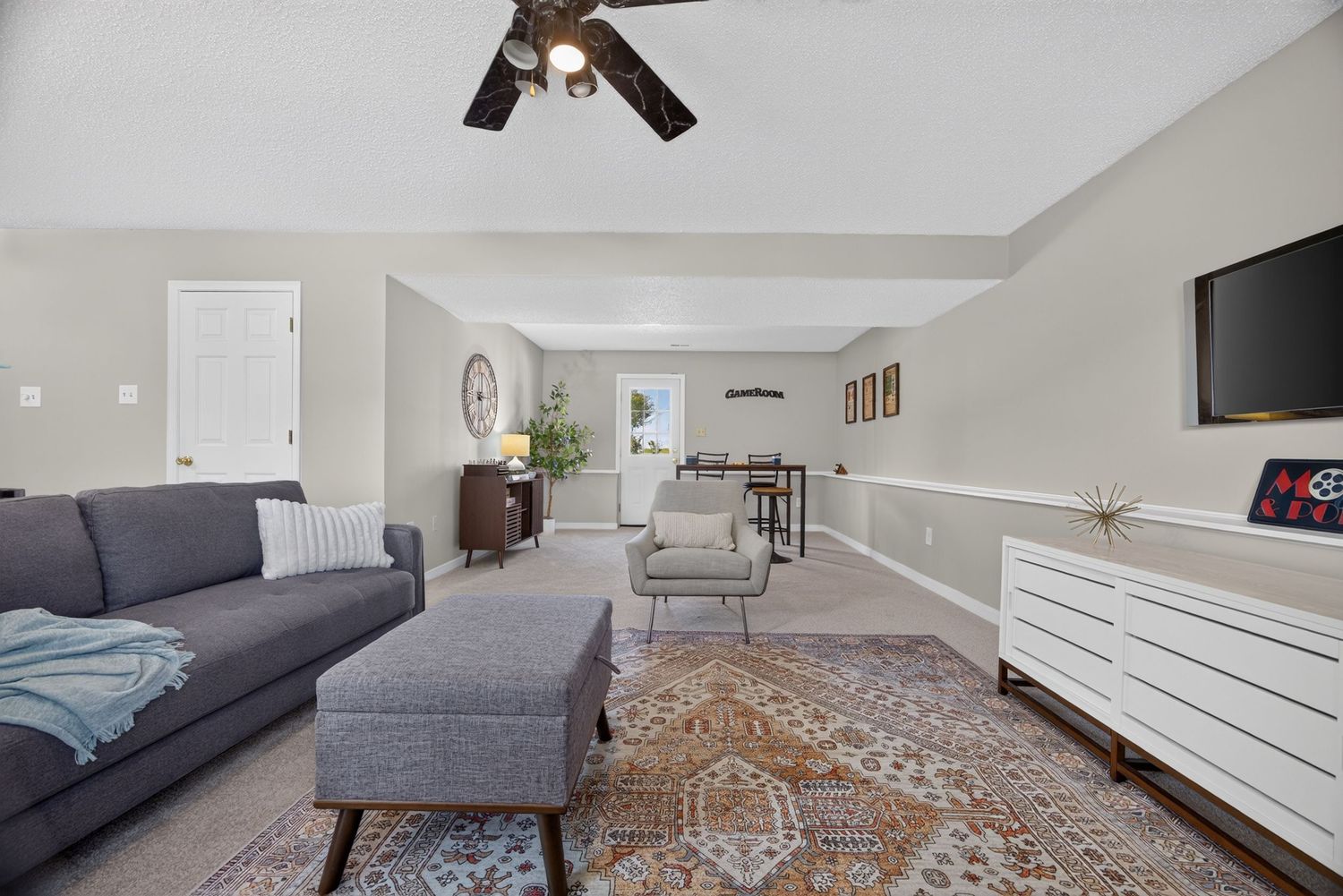 Bright living room with gray sofa, ottoman, patterned rug, ceiling fan, white dresser, and open layout with neutral walls.