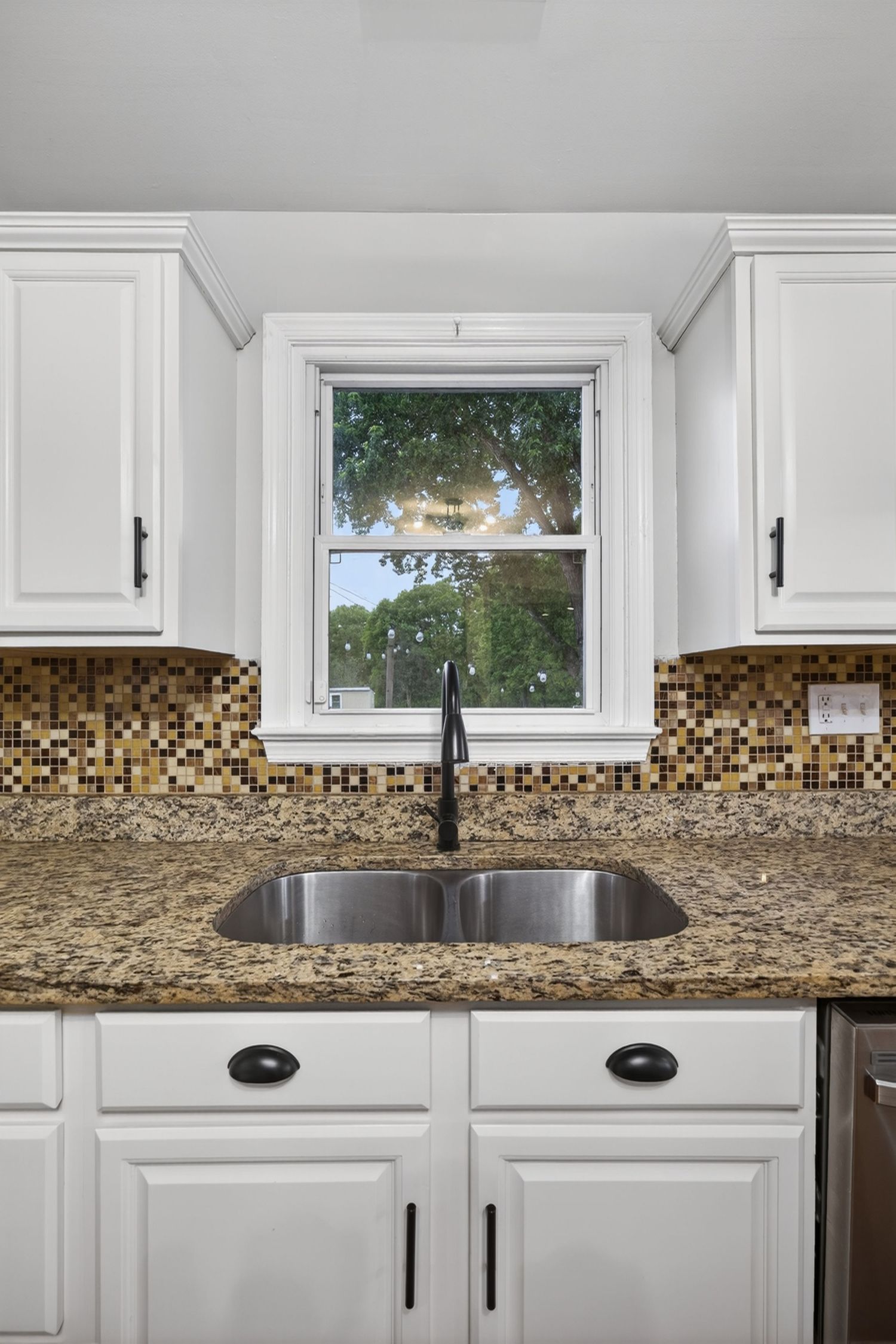 White kitchen cabinets with granite countertops, mosaic tile backsplash, stainless steel sink, and a window overlooking trees.