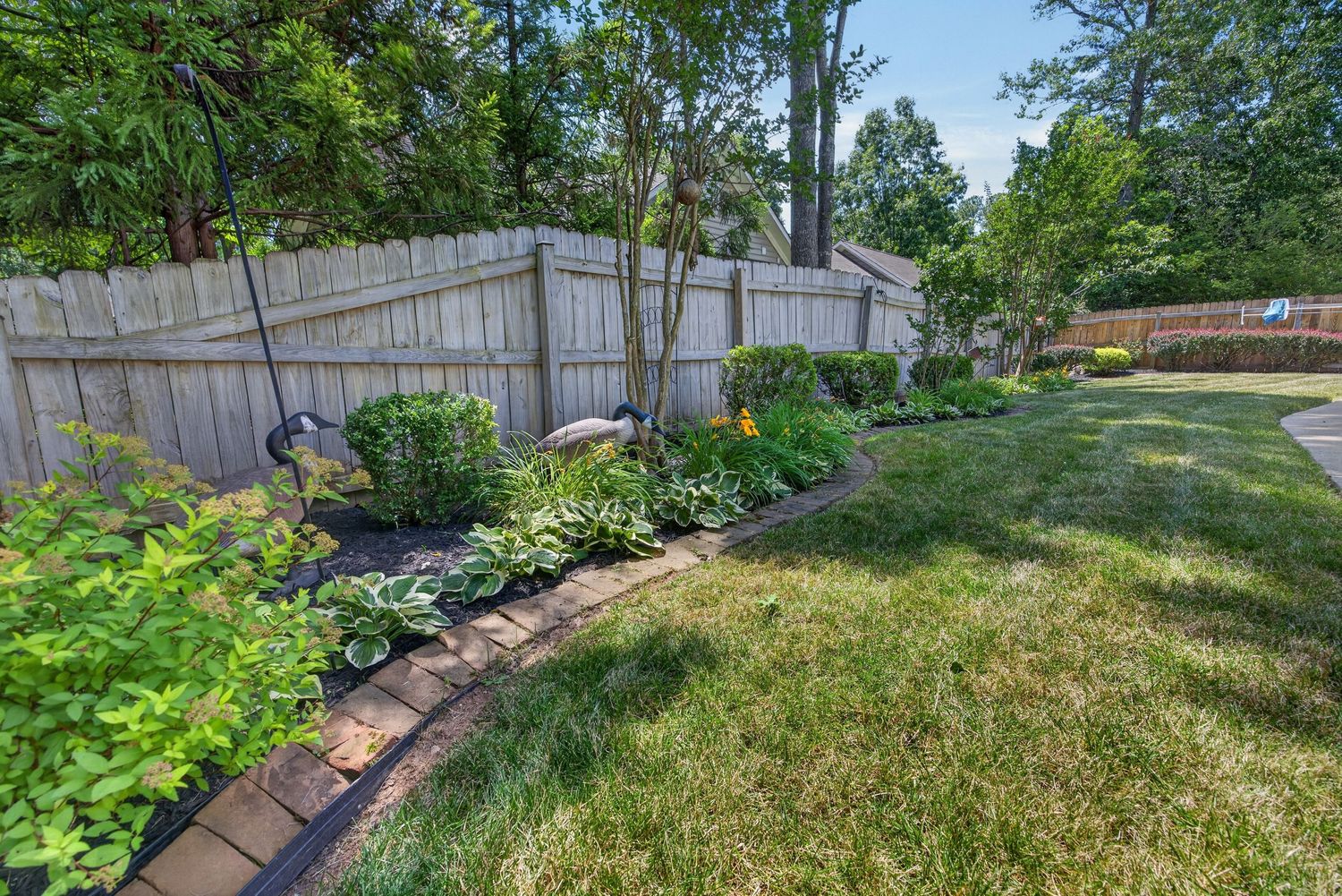 Lush backyard garden with brick edging along a wooden privacy fence, surrounded by green shrubs and a well-maintained lawn.