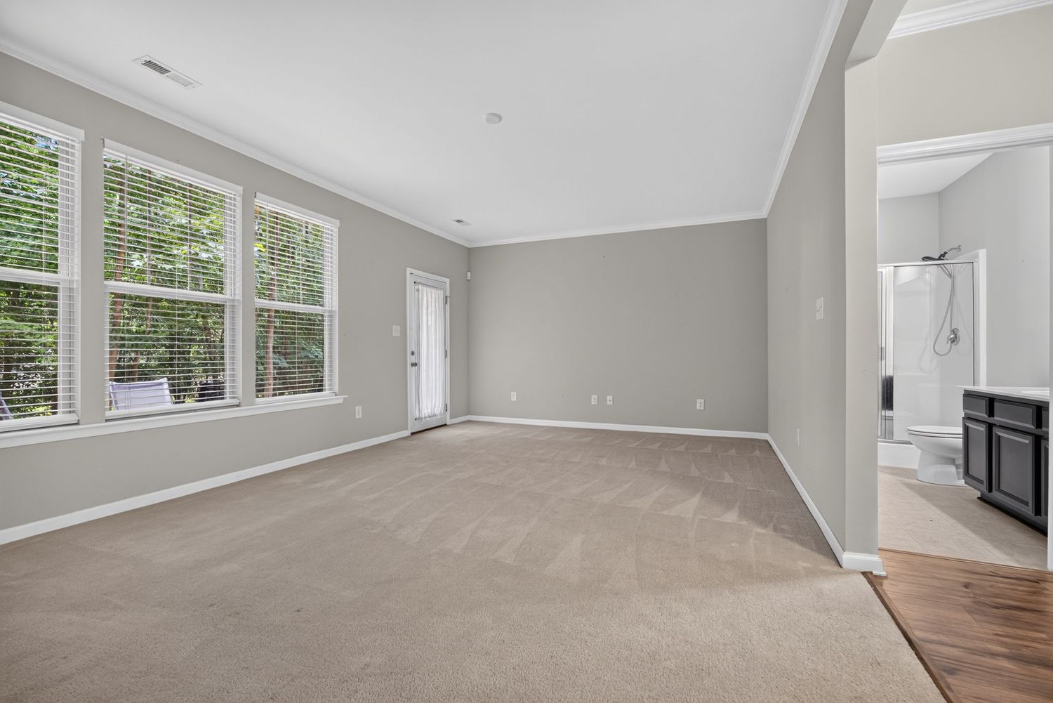 Spacious empty living room with light gray walls, crown molding, beige carpet, and large windows offering natural light.