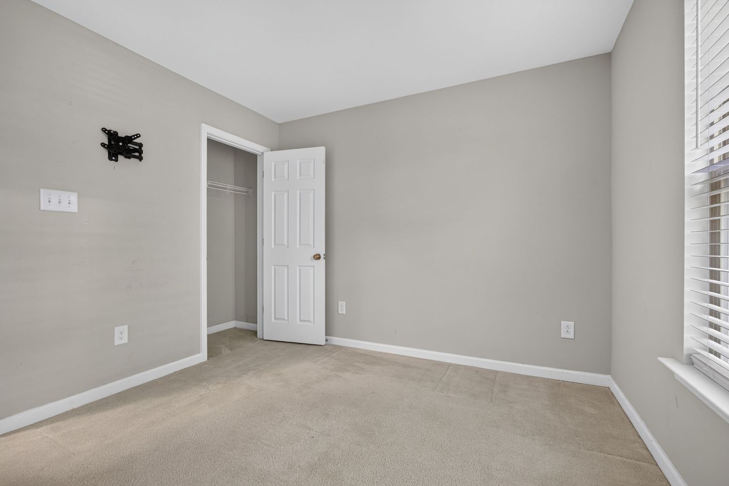 Empty bedroom with light gray walls, beige carpet, white closet door, and a window with white blinds.