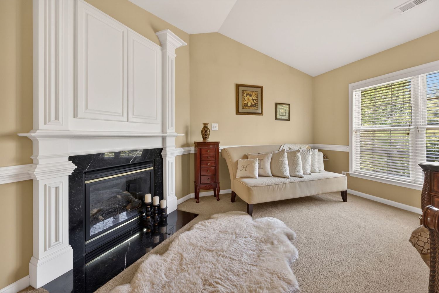 Elegant living room with white fireplace surround, black marble hearth, beige walls, carpeted floors, and a chaise lounge by a window.