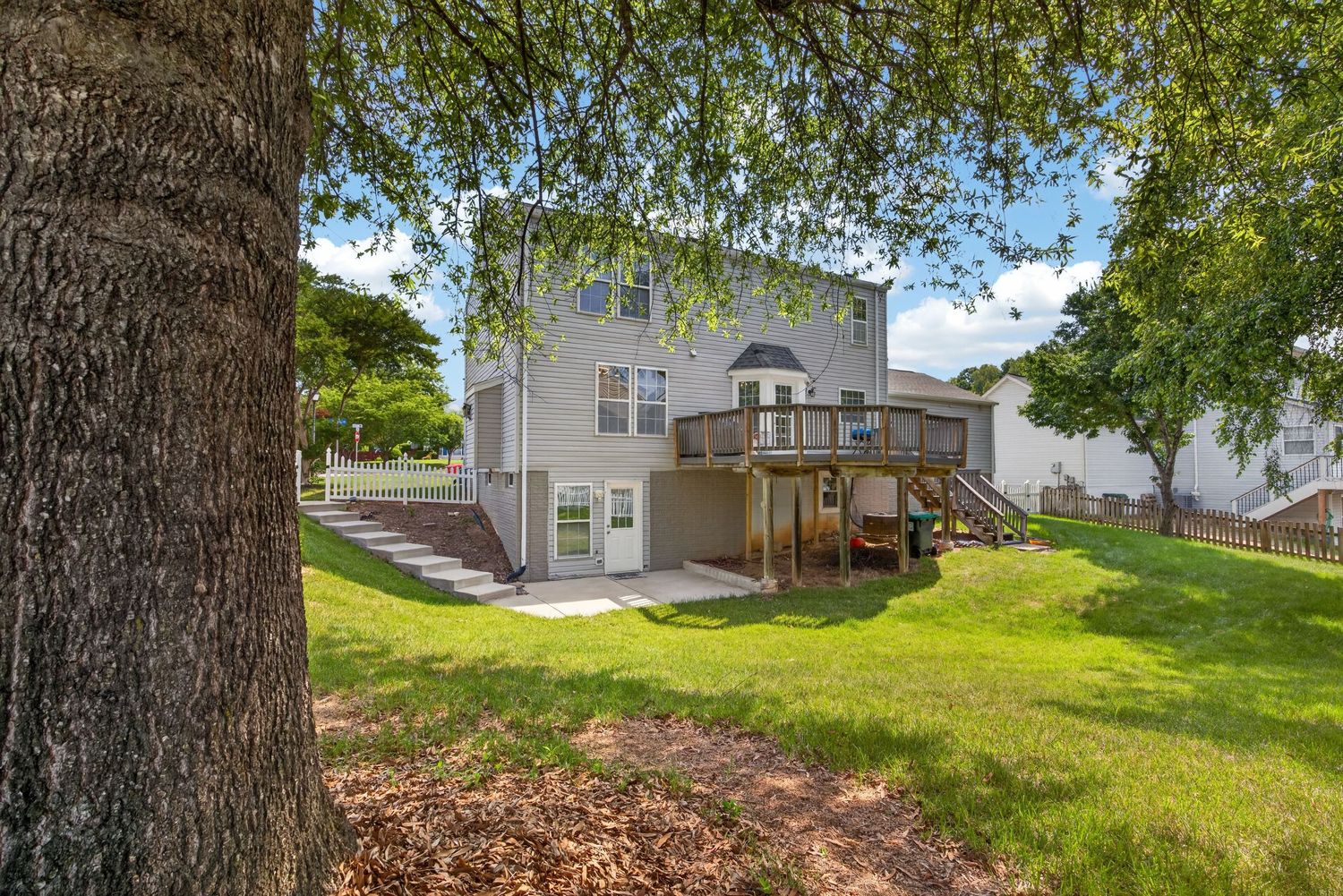 Two-story gray home with large wooden deck, lush green lawn, and mature trees in a suburban backyard setting.