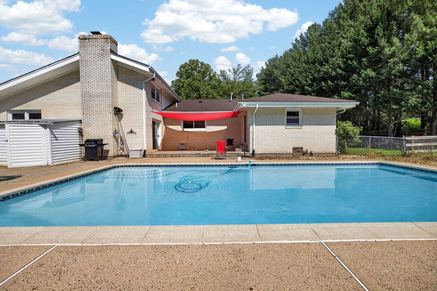 Outdoor in-ground swimming pool with red shade sail behind a brick ranch home surrounded by trees.