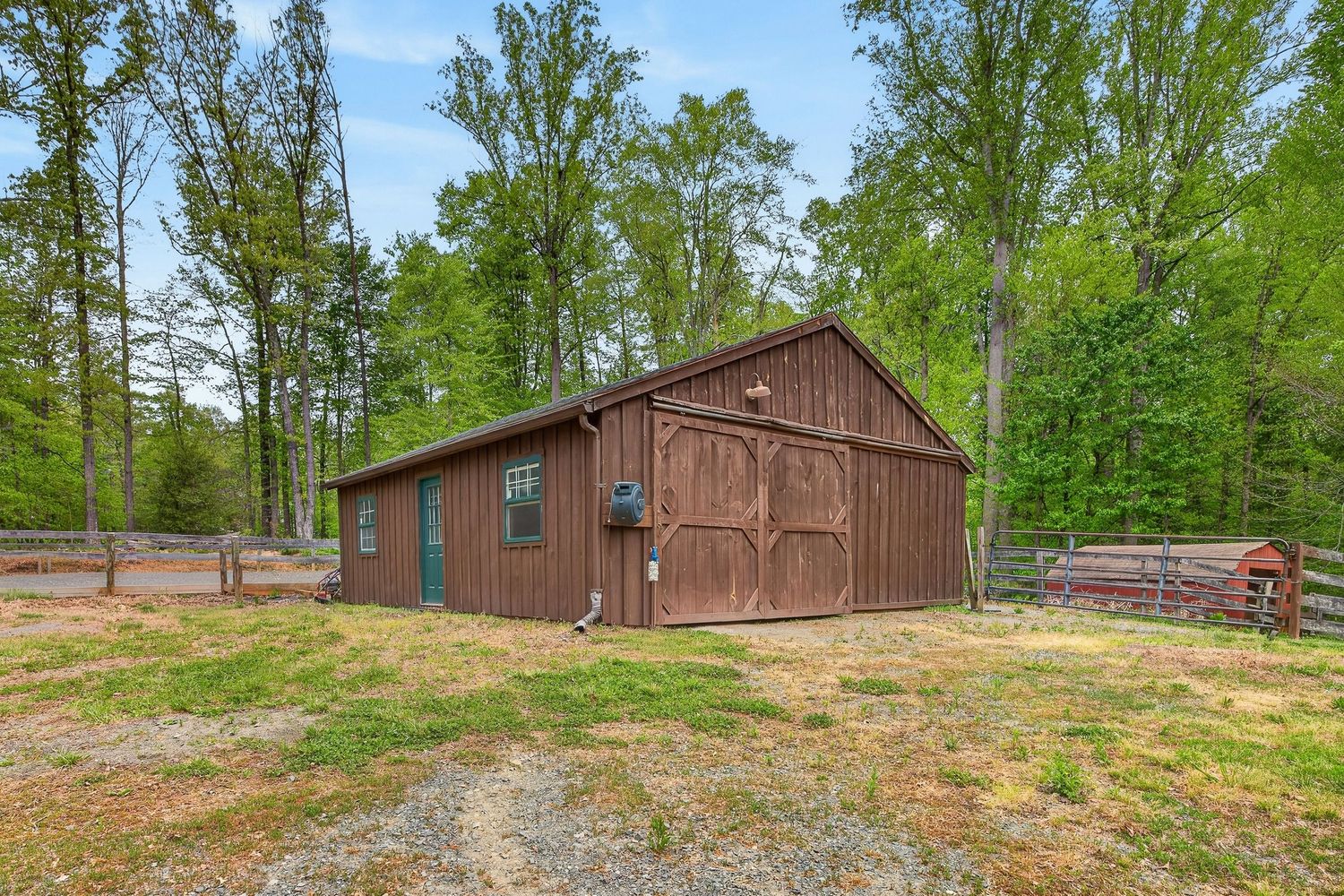 Brown barn with large wooden doors surrounded by lush green trees on a gravel lot in a rural setting.