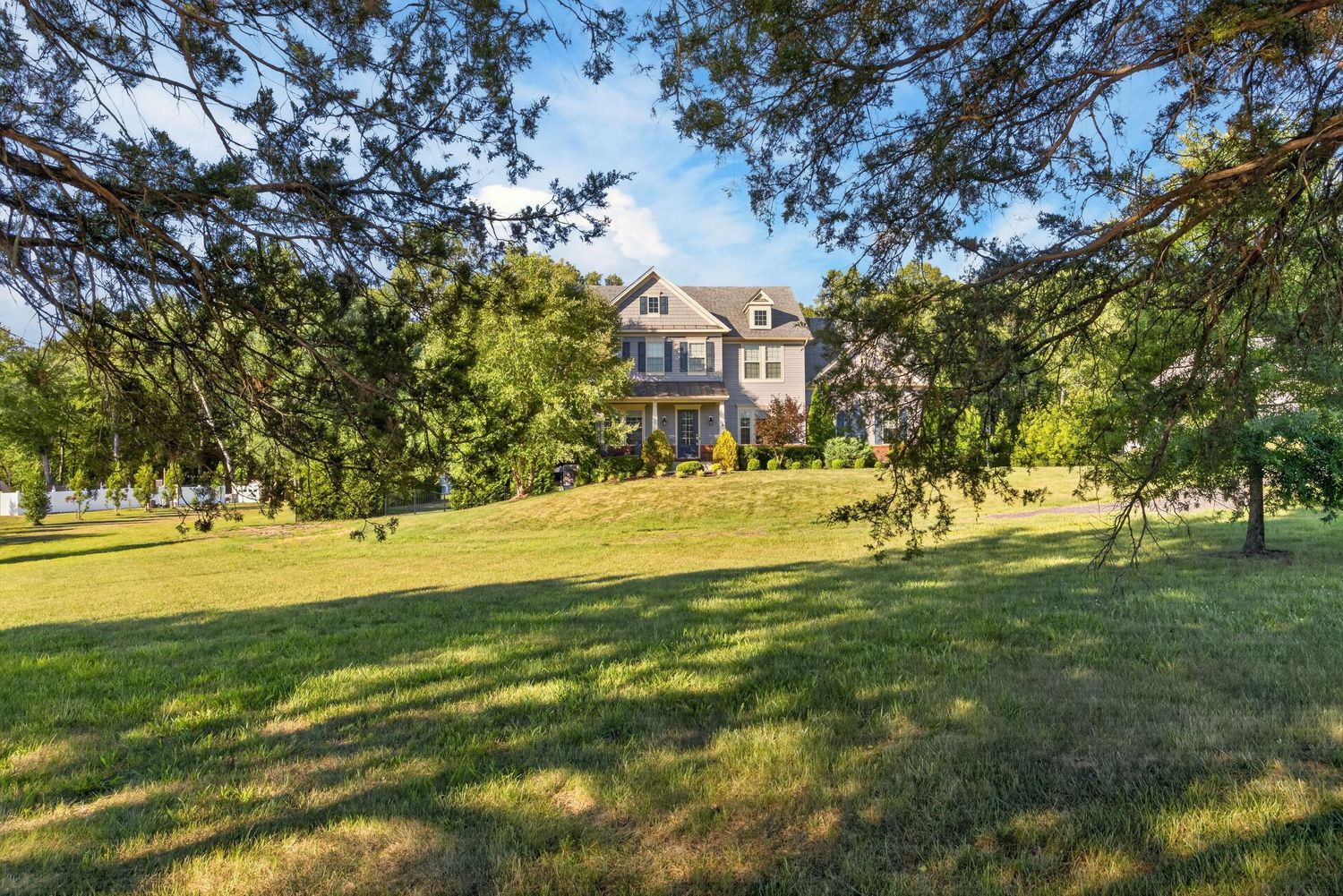 Large two-story home nestled on a lush green hillside surrounded by mature trees under a bright blue sky.