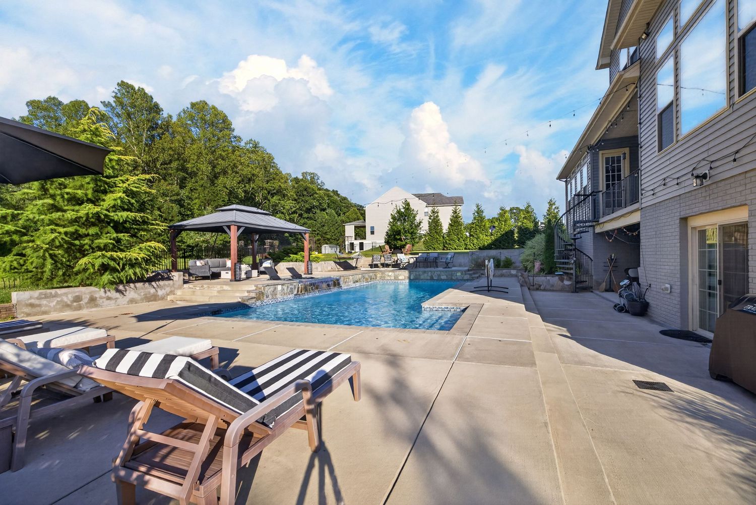 Luxury backyard pool with lounge chairs, pergola, and lush greenery on a sunny day.