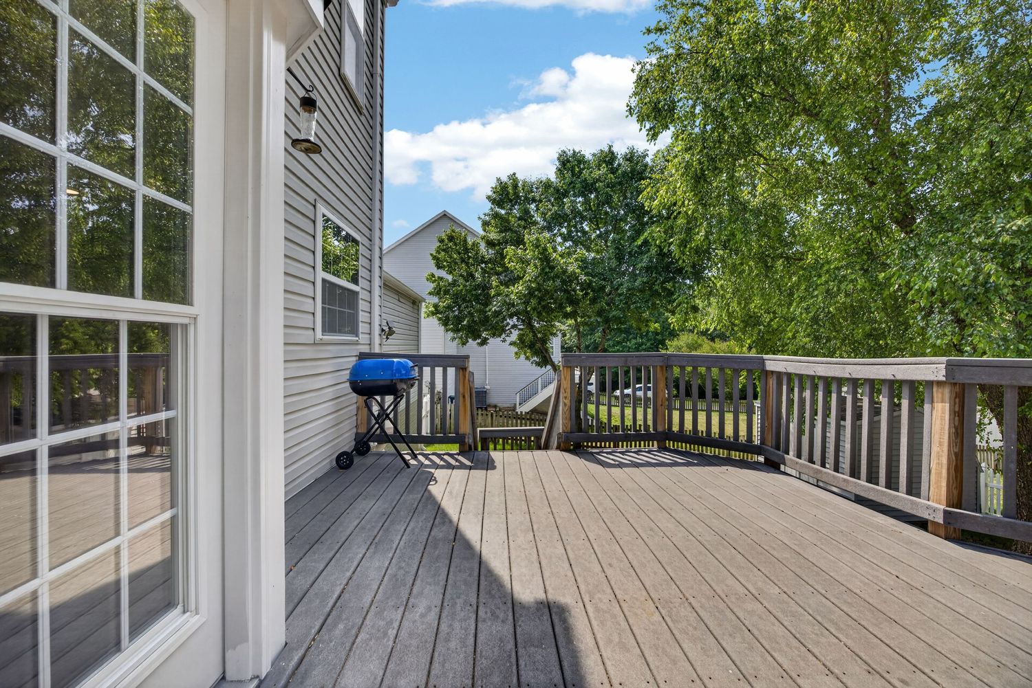 Spacious wooden deck with gas grill, wood railing, and lush green trees surrounding a suburban home.
