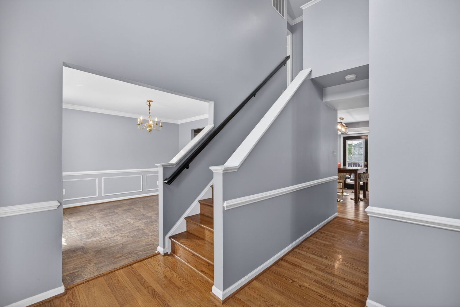 Elegant home entryway featuring hardwood floors, white wainscoting, a staircase with dark railing, and a chandelier in the background.