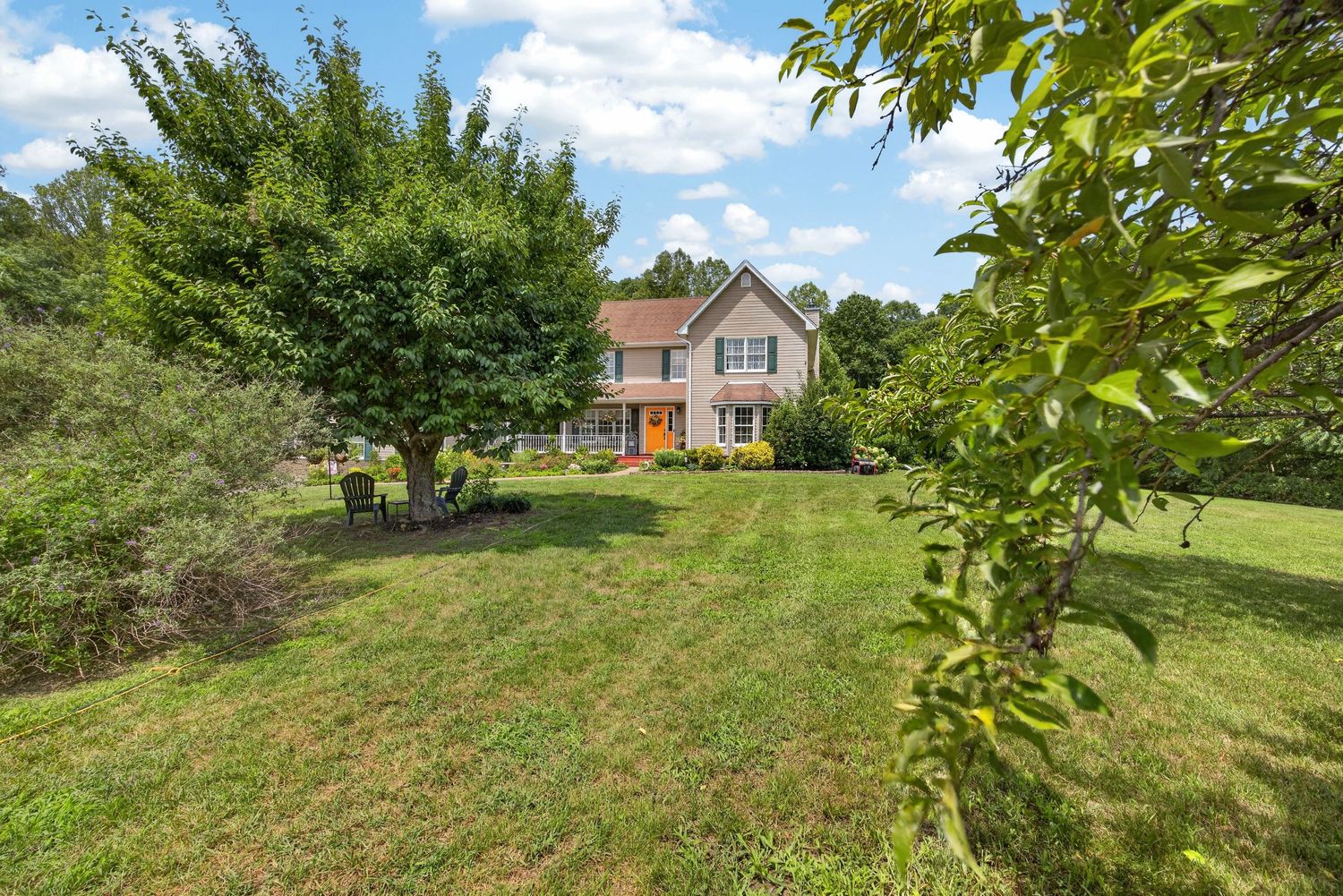 Two-story brick home with orange front door surrounded by lush green lawn and mature trees on a sunny day.