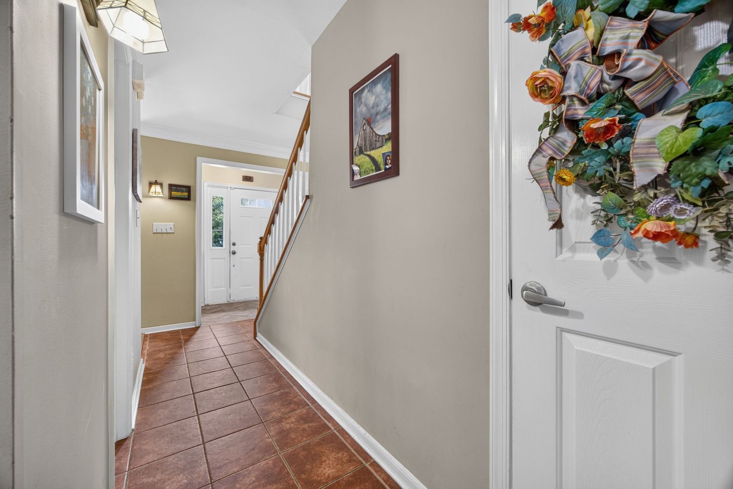 Bright entryway with terracotta tile floors, white trim, staircase, and a decorative floral wreath on the front door.