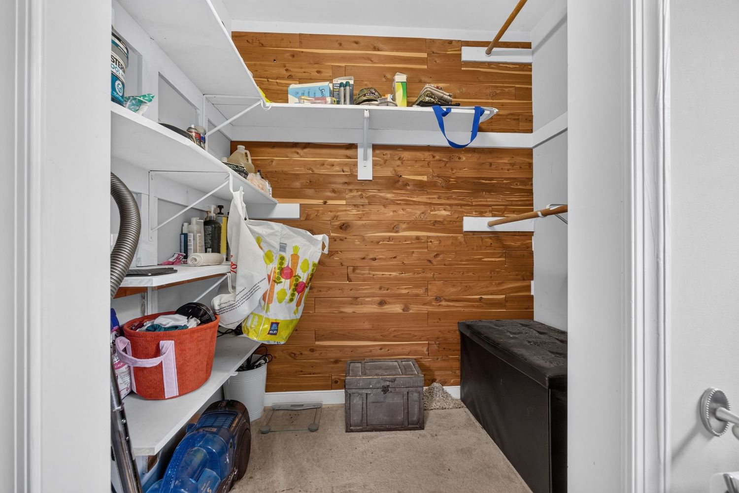 A utility room with wood-paneled walls, white shelving, washer, storage trunk, and carpeted flooring.