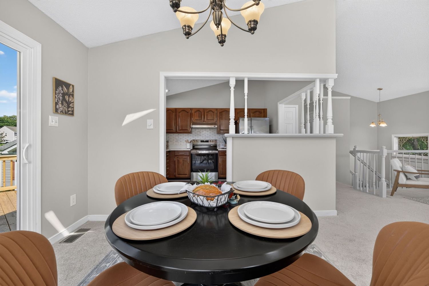 Bright dining area with round black table, brown chairs, chandelier, and open view to kitchen with vaulted ceilings.