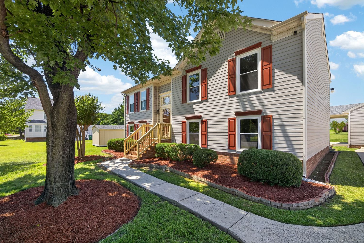 Two-story colonial home with red shutters, tan siding, brick accents, manicured shrubs, and a large shade tree on a sunny day.