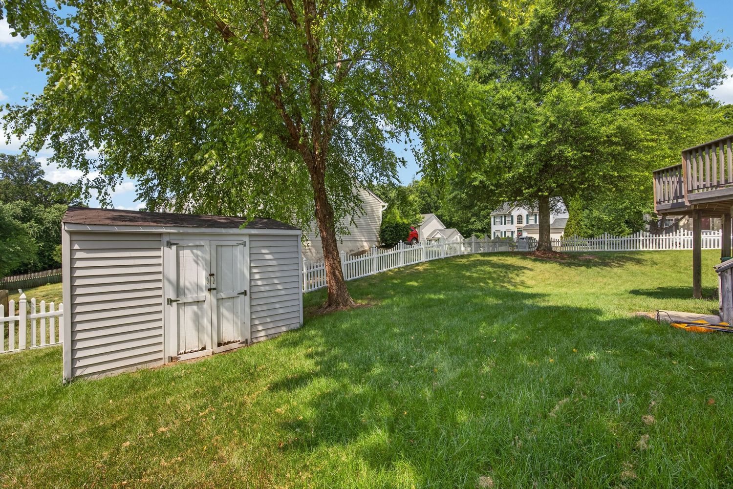 Spacious backyard with lush green lawn, storage shed, mature trees, and white picket fence on a sunny day.