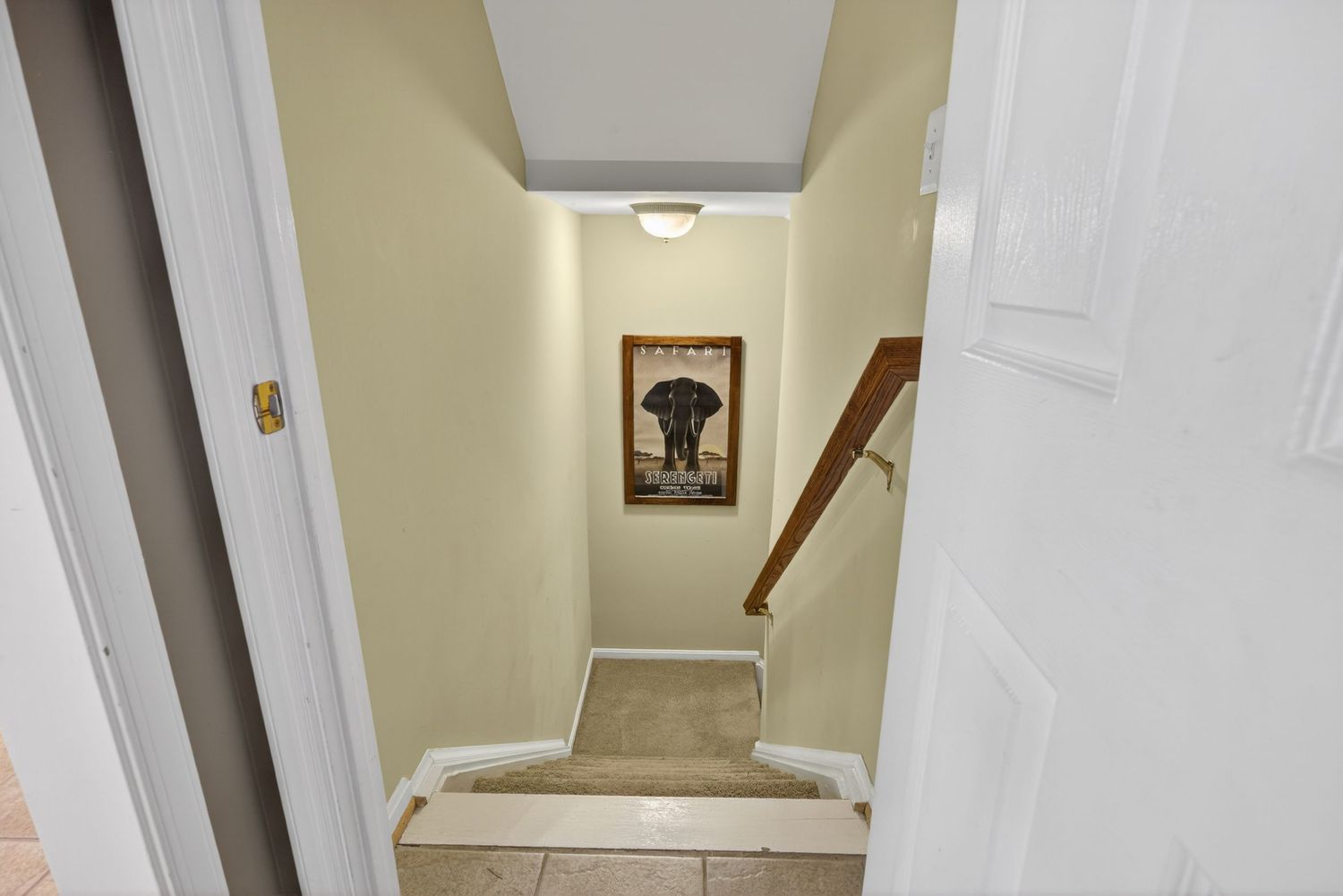 Narrow hallway with beige walls, tile flooring, carpeted staircase, and elephant artwork leading to upper level.
