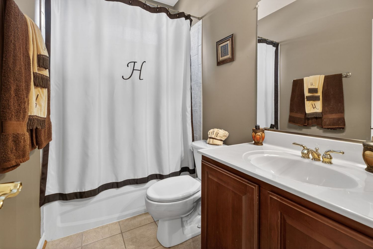 Elegant bathroom with monogrammed white shower curtain, cherry wood vanity, white sink with gold faucets, and tile flooring.
