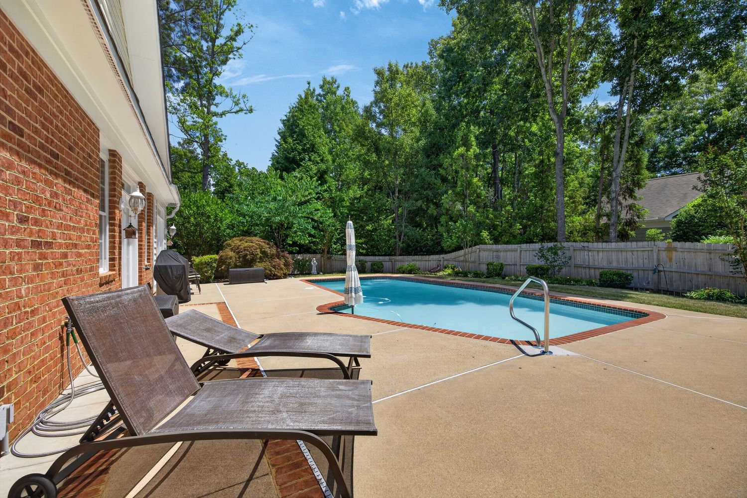 Backyard patio with lounge chairs beside an oval pool, fountain, and lush green trees behind a privacy fence.