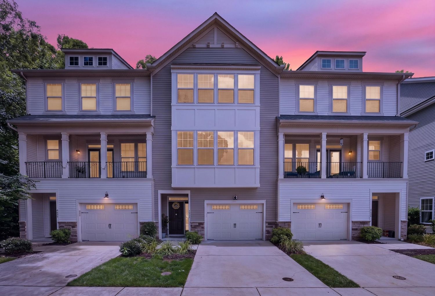 Three-story gray townhomes with garages, balconies, and large windows glow warmly against a vibrant pink and purple sunset sky.