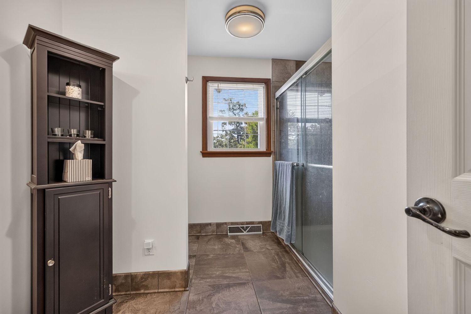 Bathroom hallway with dark wood shelving unit, glass shower door, tile floors, and a window providing natural light.