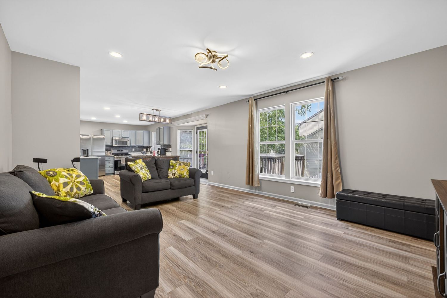 Bright open-concept living room with gray sofas, yellow accent pillows, hardwood floors, and a view into the modern kitchen.
