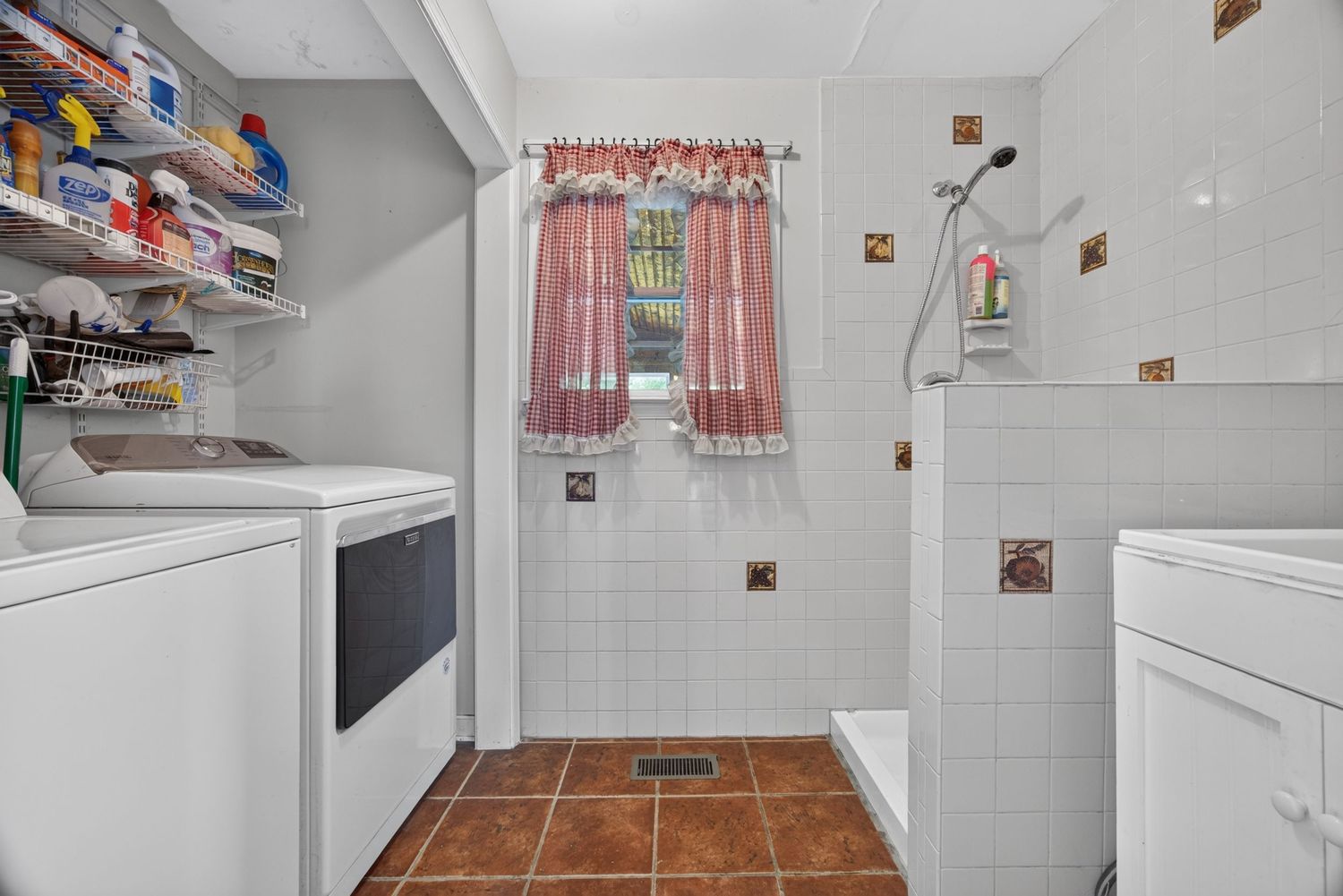 Laundry room with white washer and dryer, tiled shower, terracotta floor tiles, shelving, and a small curtained window.