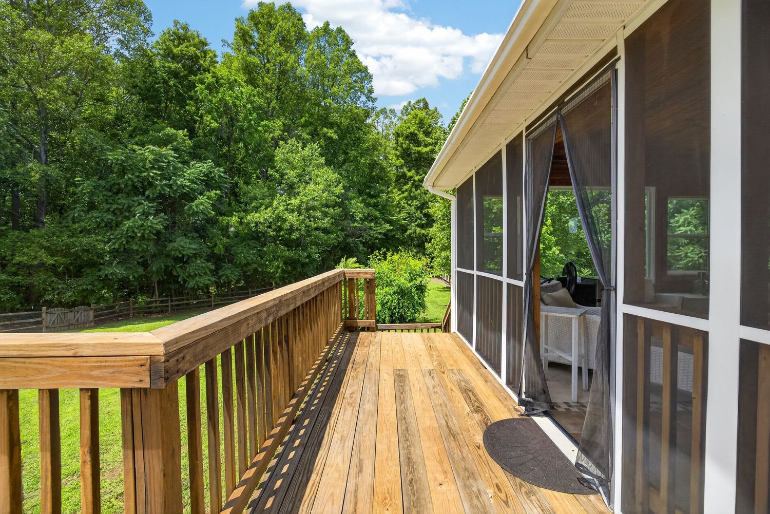 Wooden deck with white railing overlooks lush green trees, adjacent to a screened porch on a sunny day.