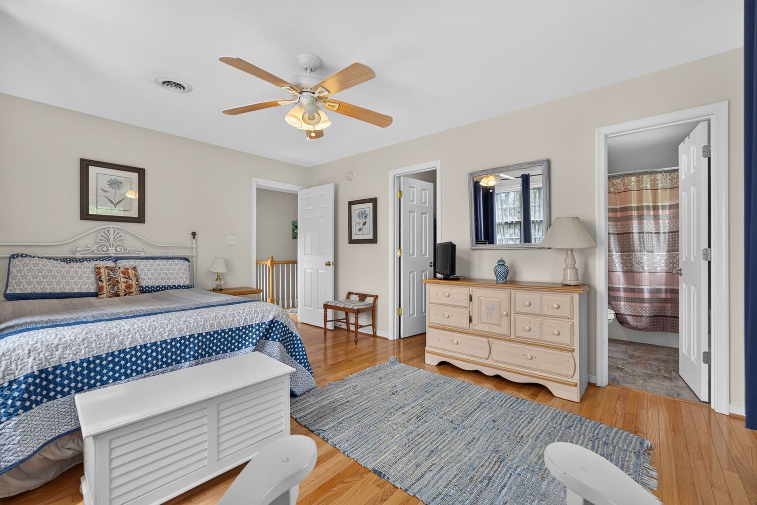 Bright bedroom with blue bedding, ceiling fan, hardwood floors, dresser, and en-suite bathroom visible through open door.