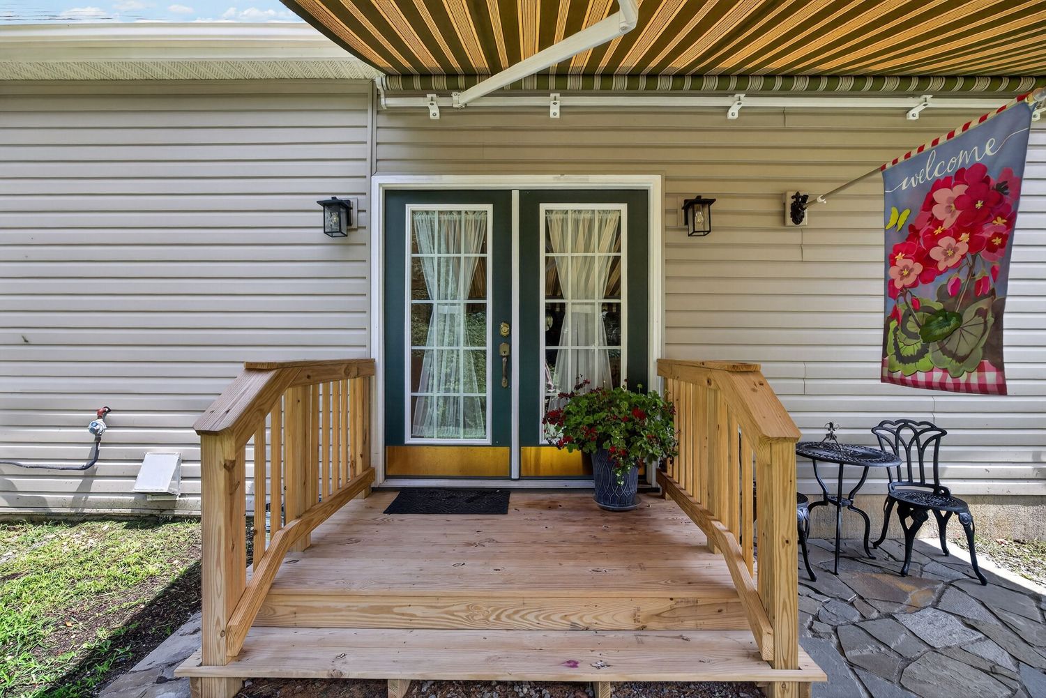 Wooden deck with green French doors, decorative wall hanging, and striped awning on a charming home exterior.