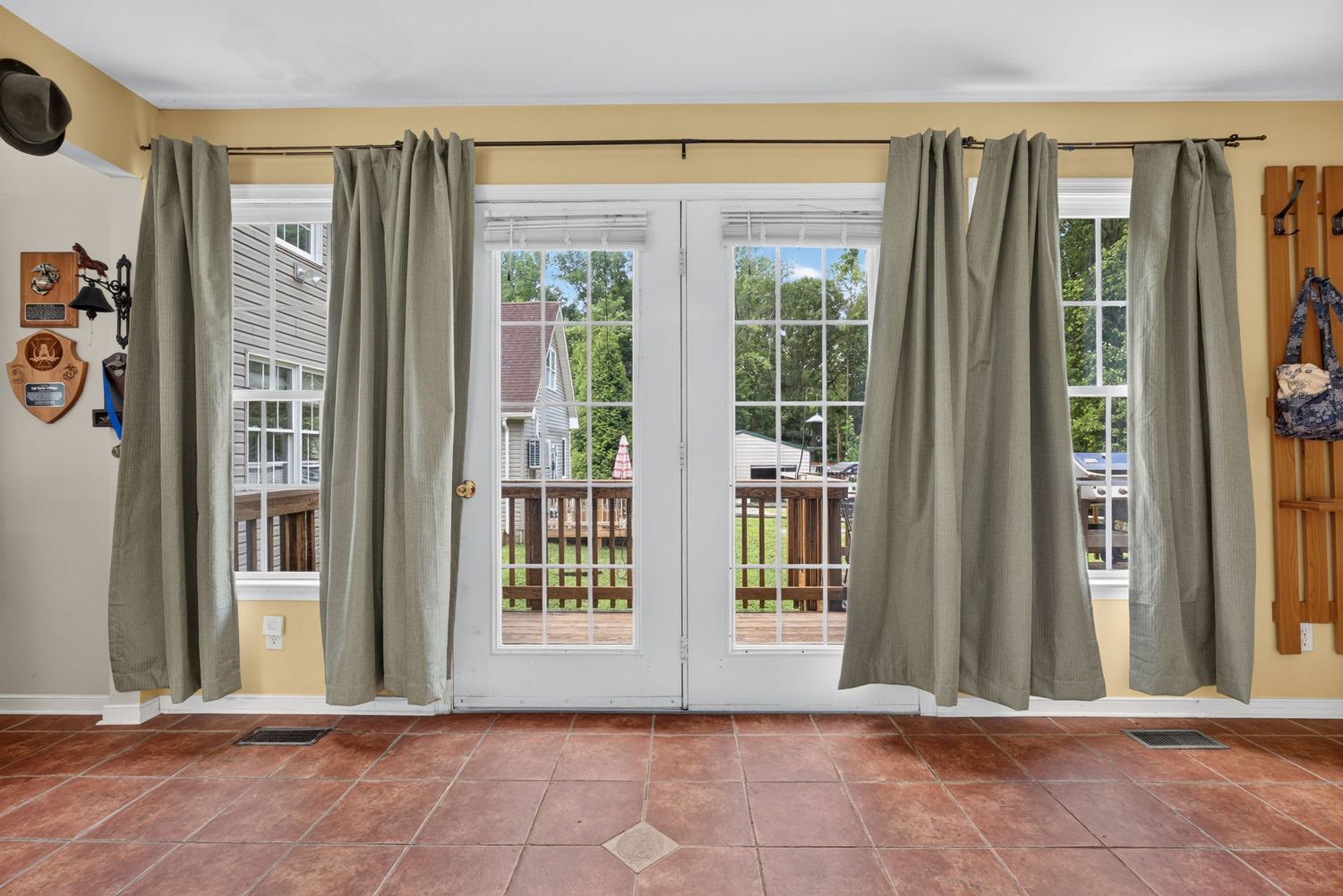 French doors with sage green curtains open to a wooden deck, featuring terracotta tile flooring in a bright home interior.