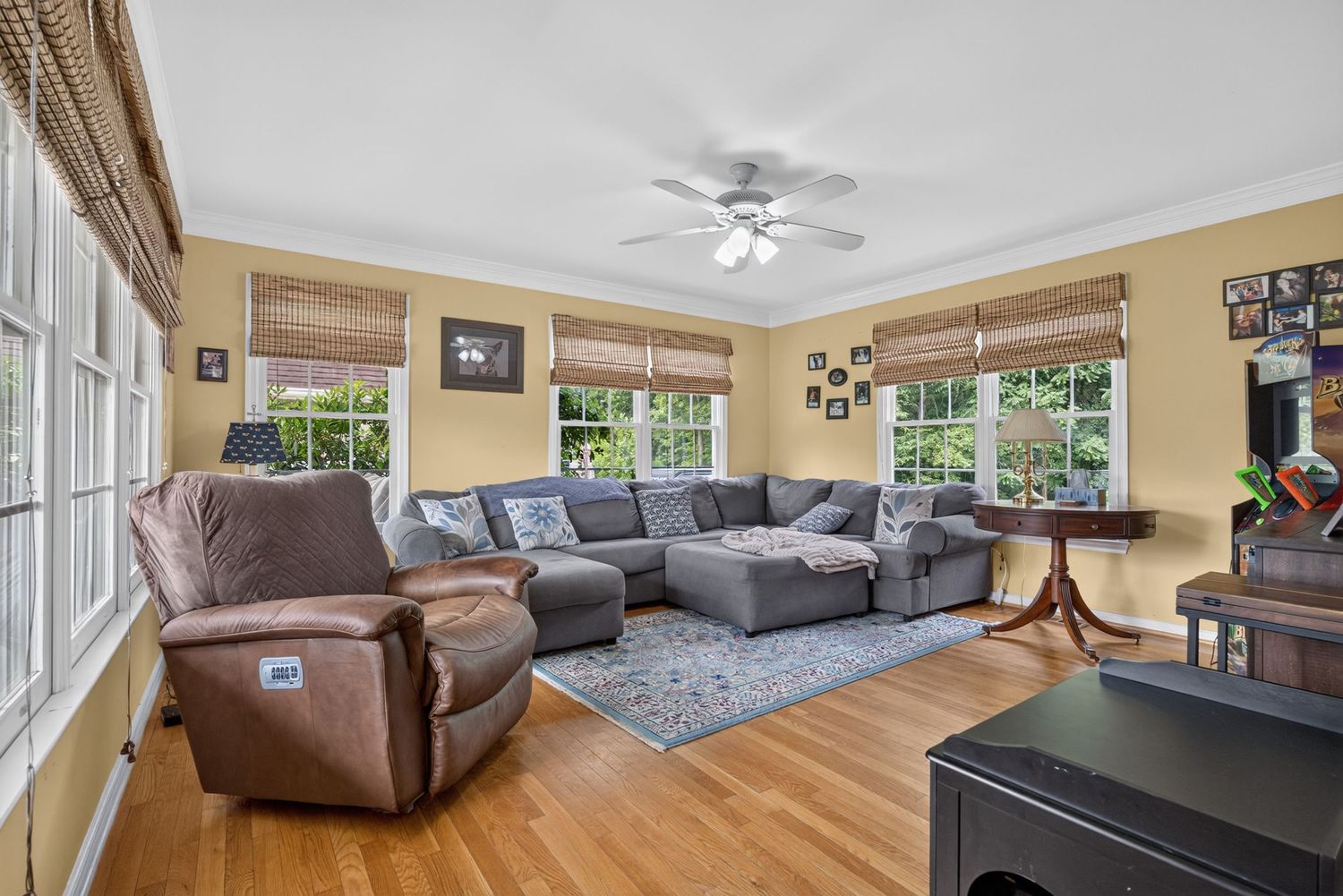 Bright living room with gray sectional sofa, recliner chair, hardwood floors, ceiling fan, and large windows with bamboo shades.