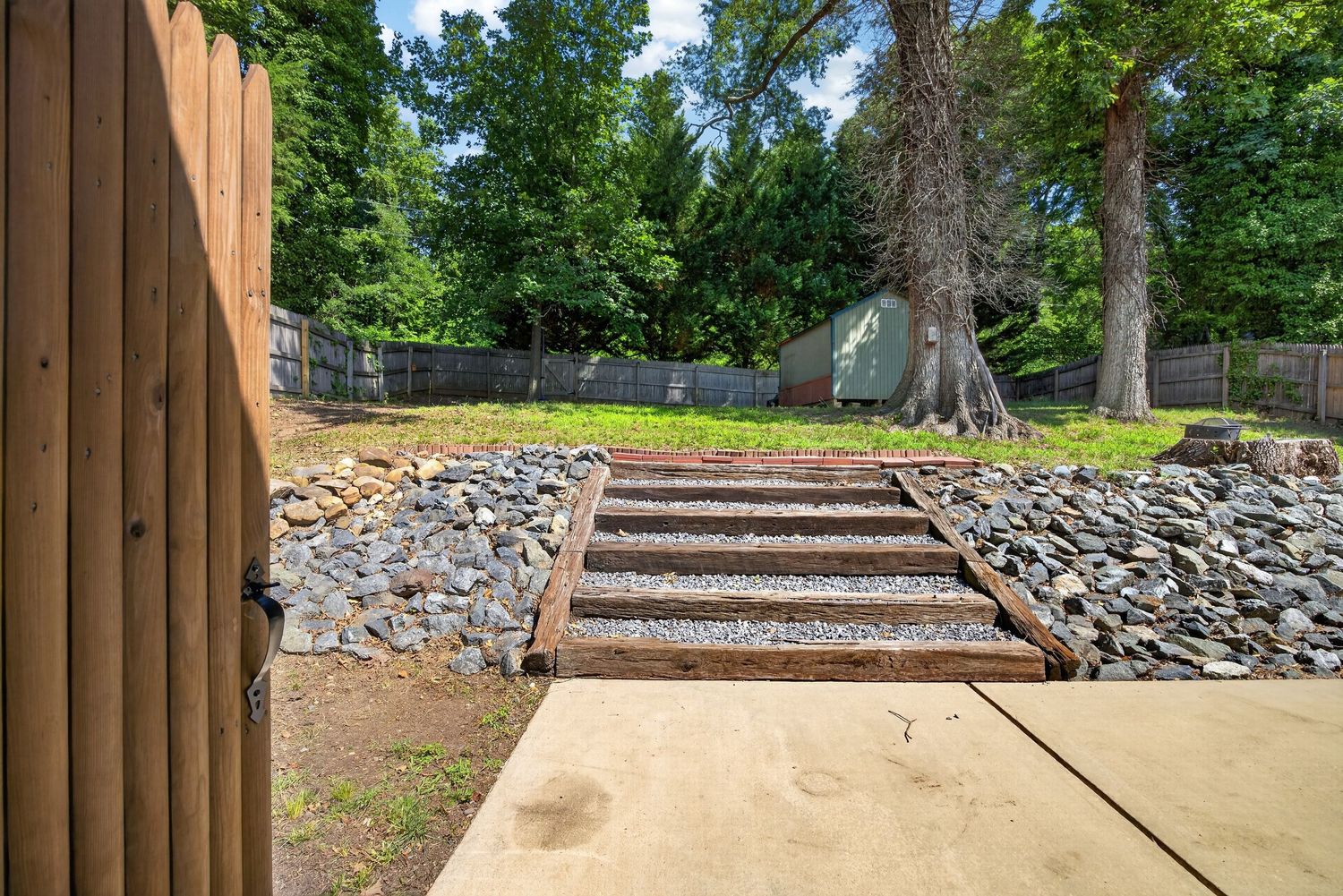 Wooden railroad tie steps with gravel landscaping lead up to a lush green backyard with mature trees and a wooden fence.