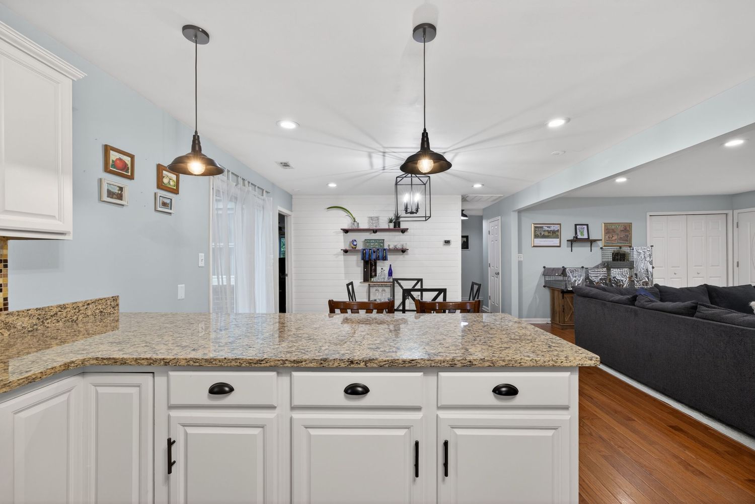 White kitchen island with granite countertop and pendant lights, open to living area with hardwood floors.