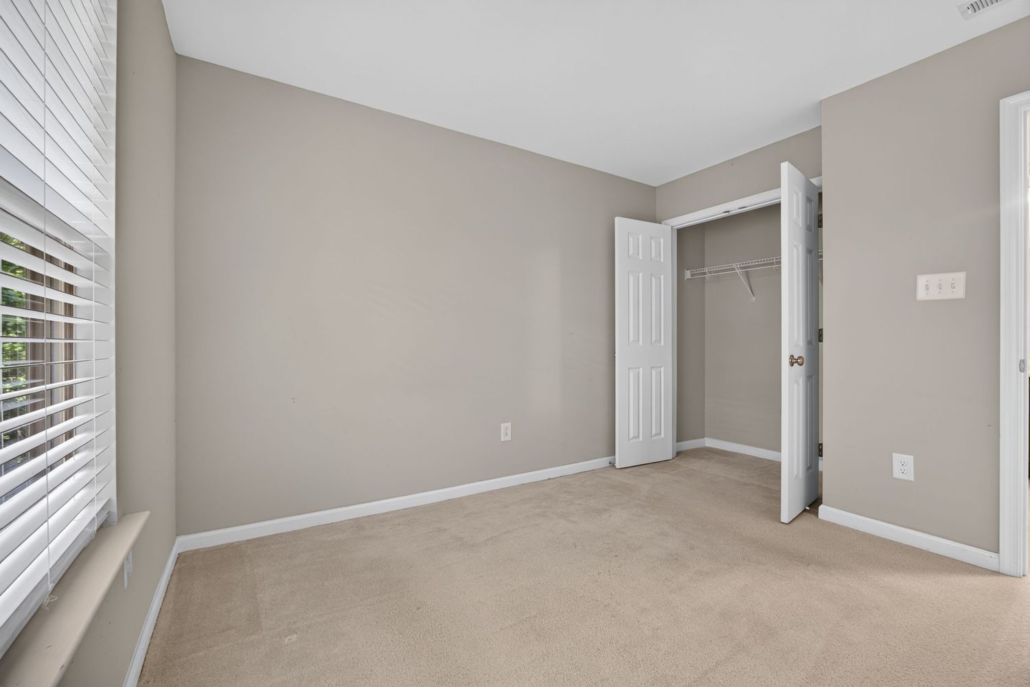 Bright empty bedroom with beige carpet, gray walls, white trim, walk-in closet, and natural light from a window.
