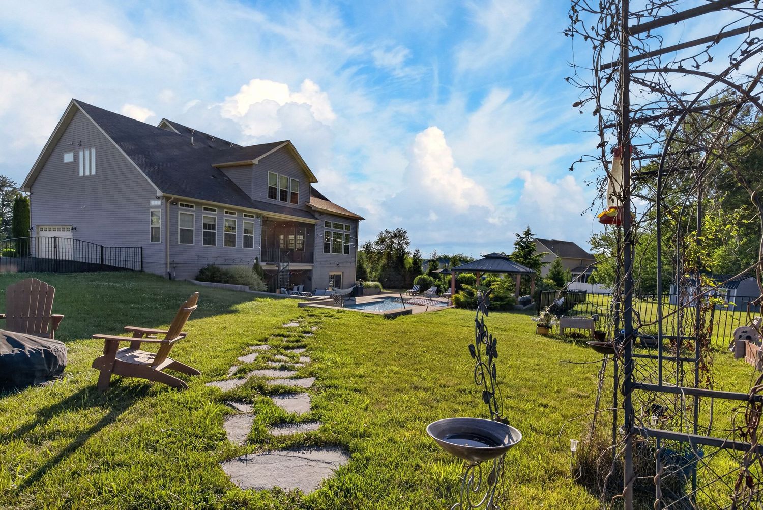 Spacious backyard with lush green lawn, stone path, Adirondack chairs, and a large two-story home under a blue sky.