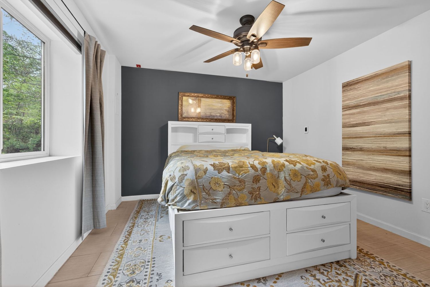 Cozy bedroom with white storage bed, gray accent wall, ceiling fan, hardwood floors, and natural light from large window.