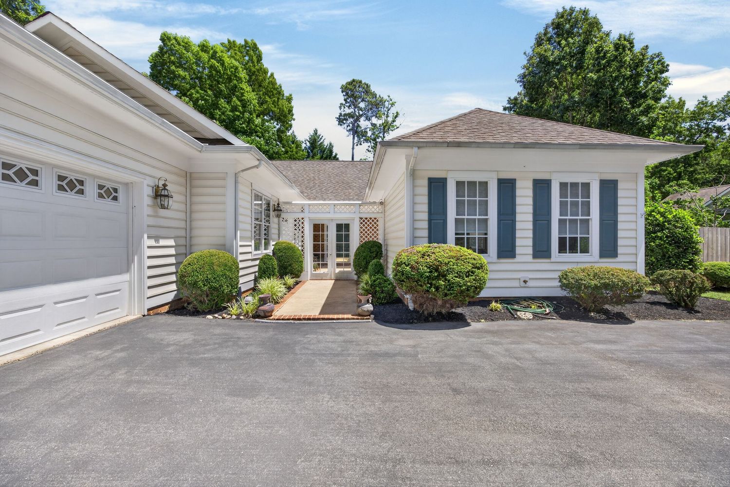 Charming white cottage-style home with blue shutters, manicured shrubs, and a two-car garage on a paved driveway.