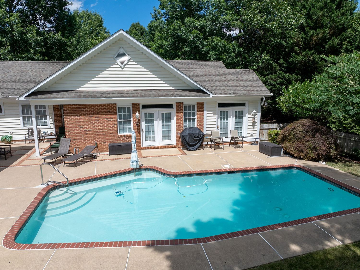 Outdoor swimming pool with brick patio, lounge chairs, and a charming house surrounded by lush green trees.