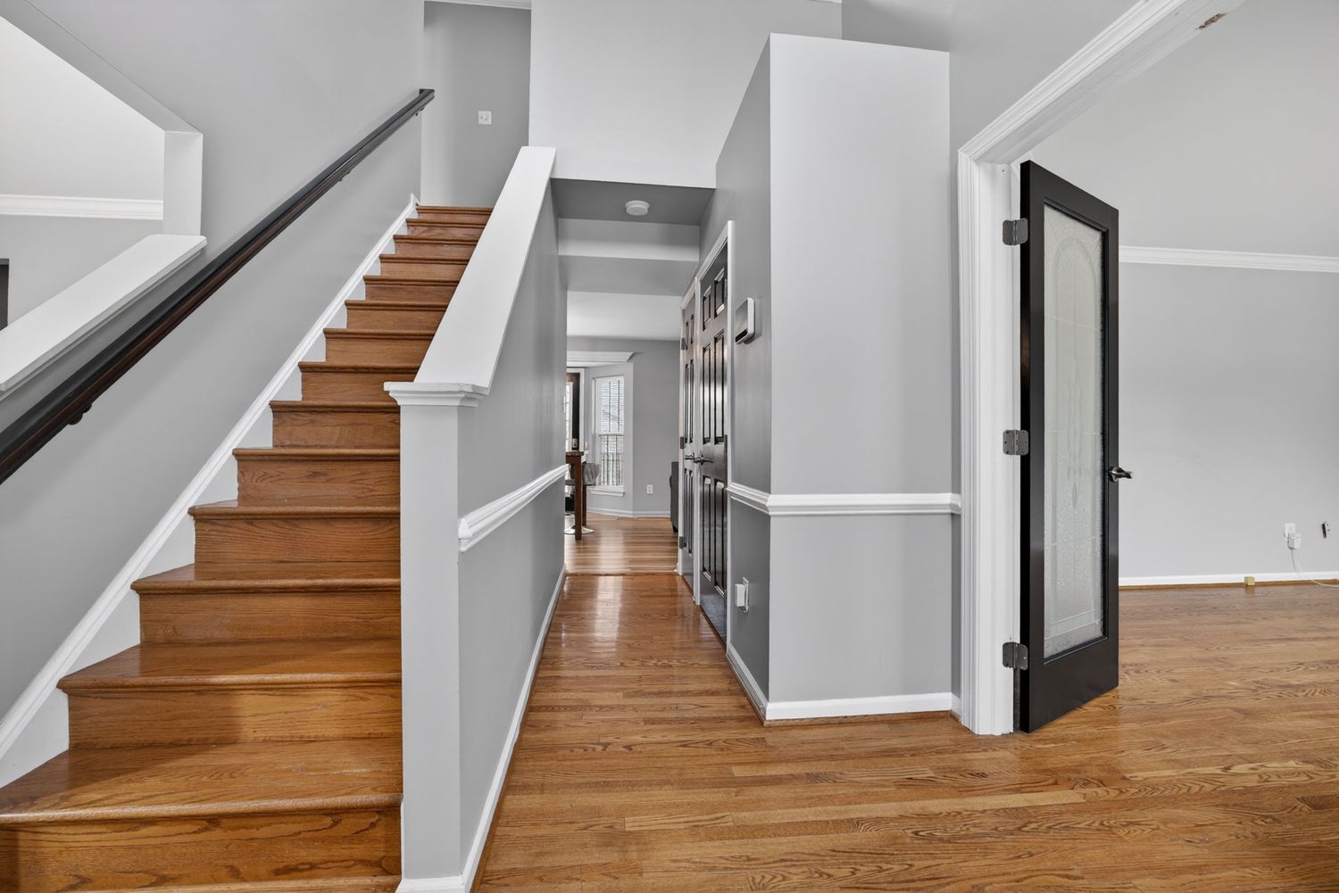 Bright hallway with hardwood floors, white walls, staircase with wood steps, and a black-framed glass door.
