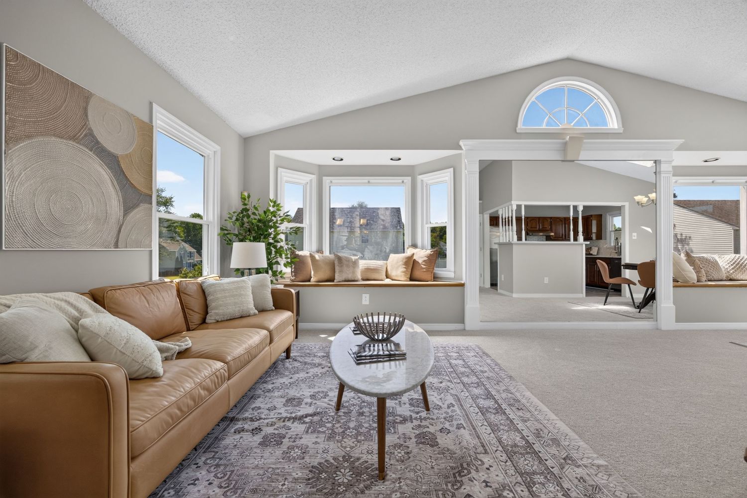 Bright living room with vaulted ceilings, tan leather sofa, round coffee table, plush carpet, and bay window with natural light.