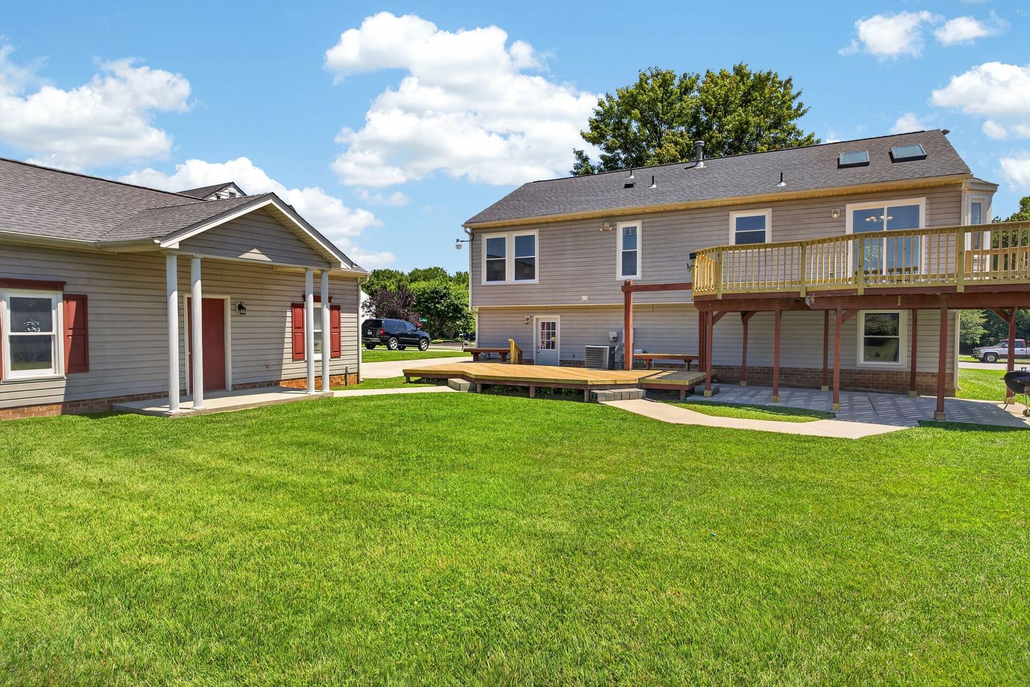Two-story residential home with large deck, attached single-story wing, and well-maintained green lawn on a sunny day.