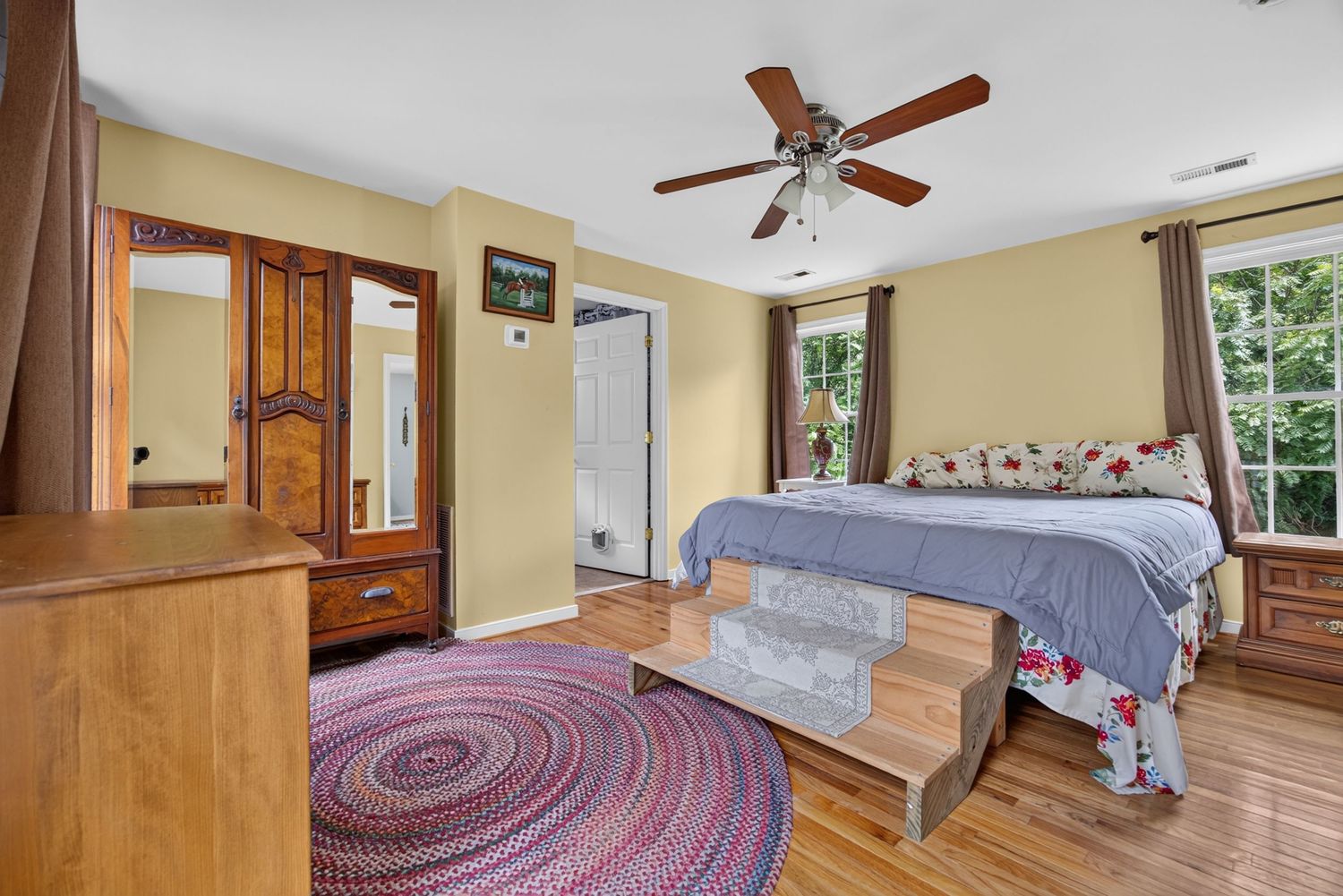 Bright bedroom with hardwood floors, ceiling fan, wood furniture, braided rug, and large windows providing natural light.