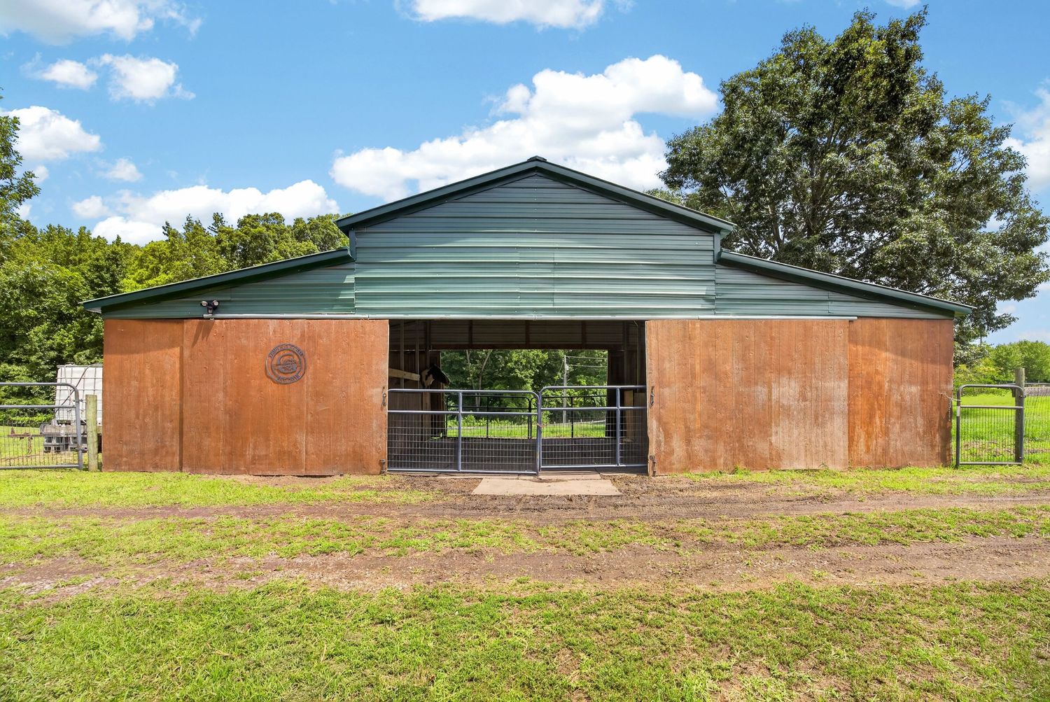 A rustic metal barn with green roof and open gates surrounded by lush green pasture and trees under a blue sky.