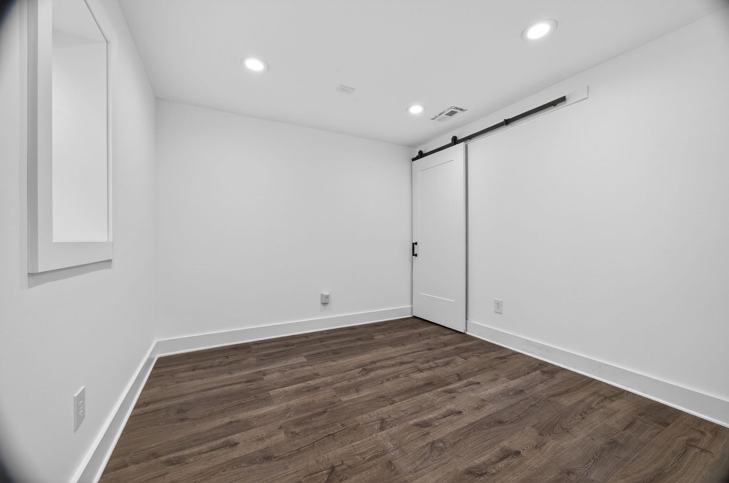 Bright empty room with white walls, dark hardwood floors, recessed lighting, and a modern barn door.