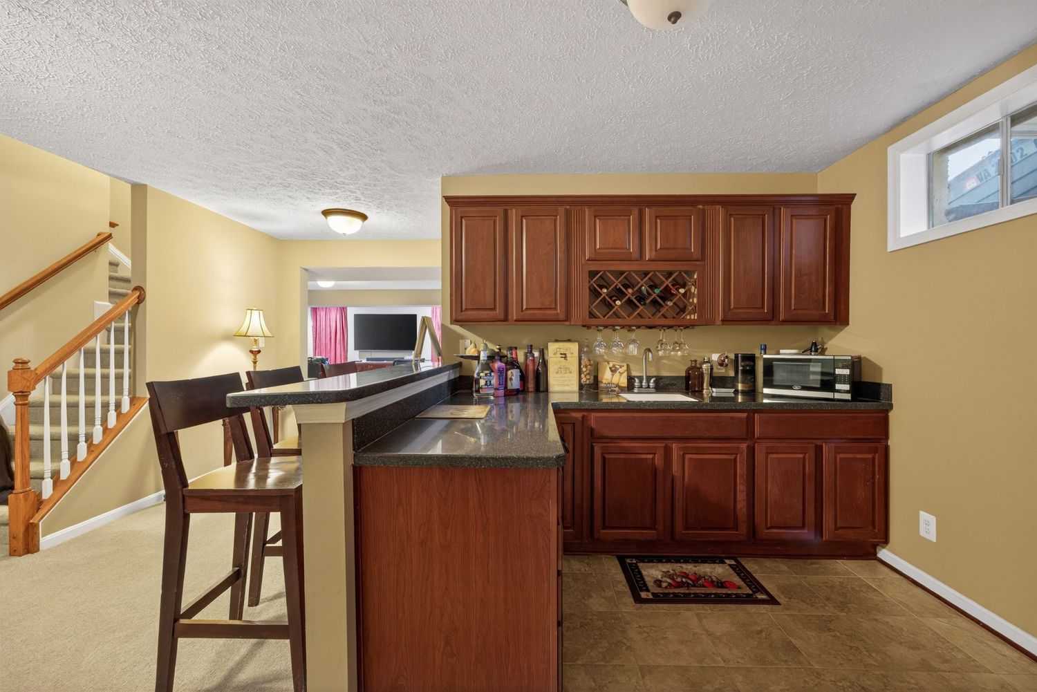Basement wet bar with cherry wood cabinets, dark granite countertop, bar seating, and wine rack near staircase.