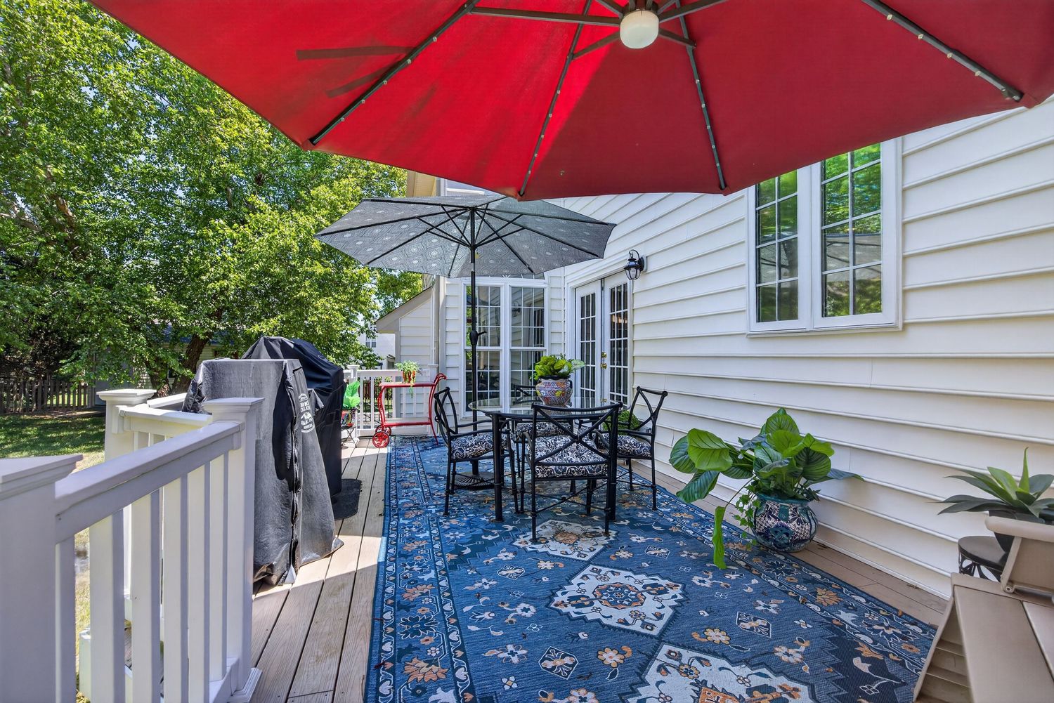 Outdoor deck with blue patterned rug, dining set under red umbrella, potted plants, and white siding home exterior.