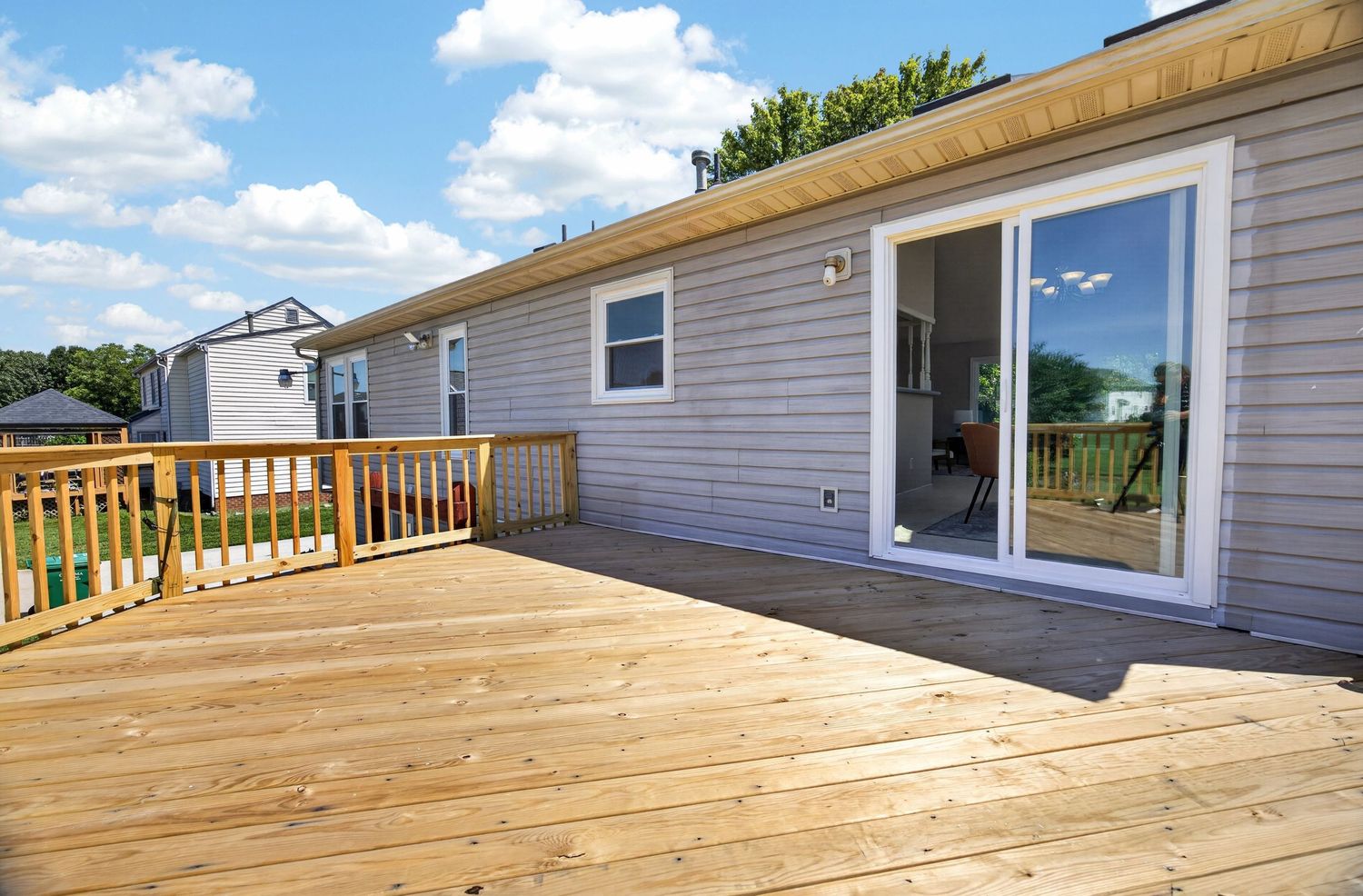 Spacious wooden deck attached to a gray vinyl siding home with sliding glass door and railing under a blue sky.